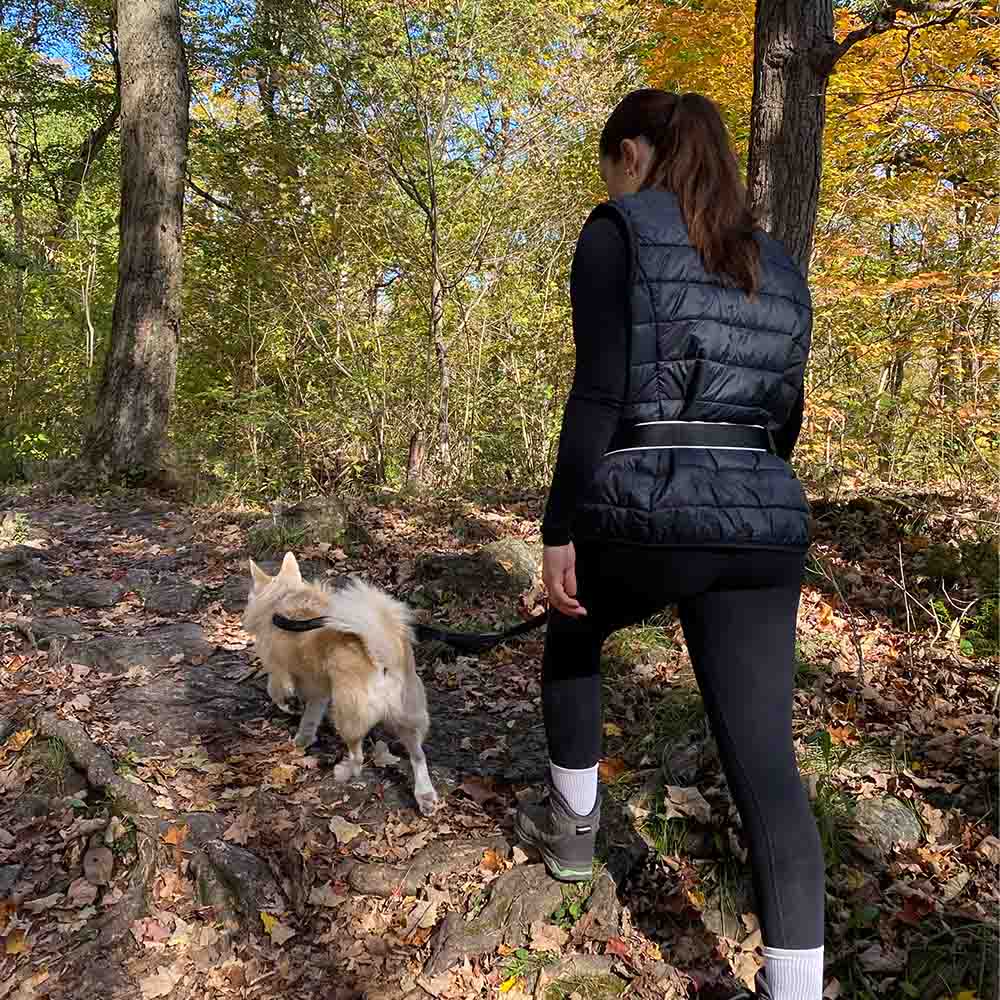 Woman hiking through forest trail using a hands-free dog leash, keeping her pet close while enjoying a safe outdoor walk.