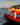Small dog wearing a bright orange and yellow life jacket standing on a red canoe, ready for safe water activities and swimming.